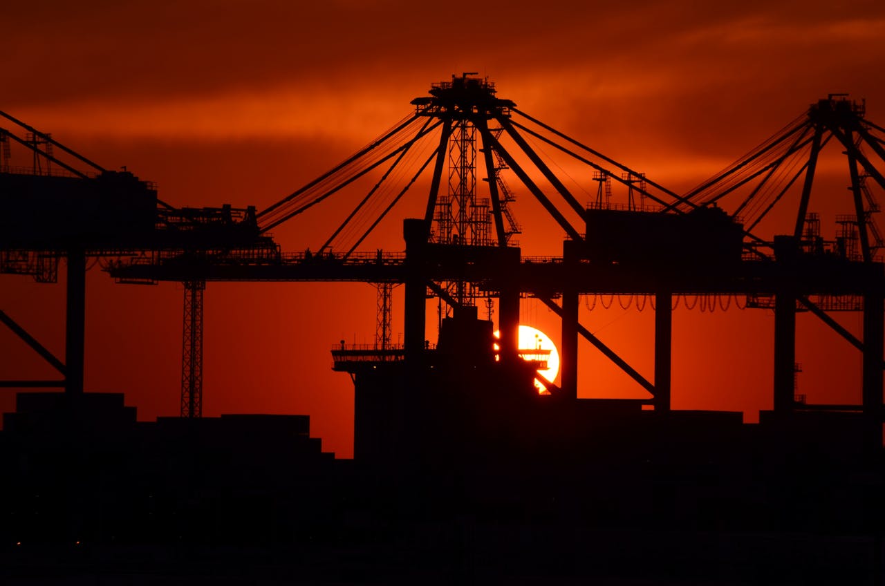 Silhouette of cargo cranes against a vivid sunset at an industrial container port.